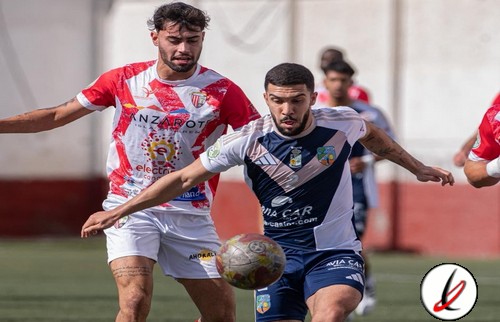 Un jugador del equipo con camiseta blanca y azul lucha por el balón con un rival con camiseta roja en un partido de fútbol en el campo de Lanzarote Deportiva.