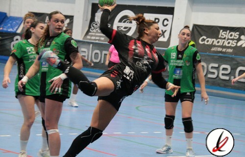 Una jugadora de balonmano en acción, saltando para lanzar el balón durante un partido femenino en un pabellón deportivo. Jugadoras en uniforme verde y negro en el fondo, en un partido de deportes en Lanzarote.
