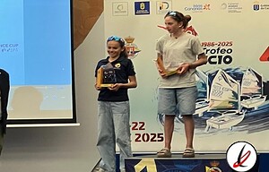 Una niña y una joven en un podio recibiendo premios en una ceremonia deportiva, con banderas y logotipos relacionados con el deporte en el fondo.
