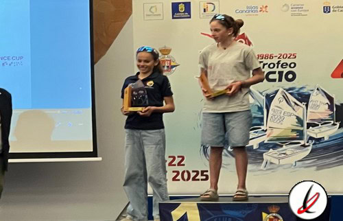 Una niña y una joven en un podio recibiendo premios en una ceremonia deportiva, con banderas y logotipos relacionados con el deporte en el fondo.