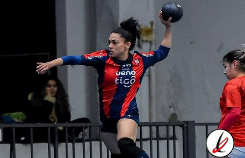 Arbusera feminina de balonmano en acción durante un partido en un pabellón cubierto, con jugadora en movimiento lanzando un balón, mostrando concentración y esfuerzo.