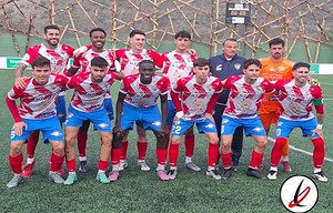Jugadores del equipo de fútbol en uniforme rojo y azul, posando en el campo de futbol antes de un partido, con un entrenador al fondo y un fondo de malla protectora en el estadio.