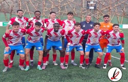 Jugadores del equipo de fútbol en uniforme rojo y azul, posando en el campo de futbol antes de un partido, con un entrenador al fondo y un fondo de malla protectora en el estadio.