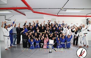 Entrenadores y niños en un dojo de judo, celebrando con una copa y medallas tras una competición, en un ambiente de alegría y triunfo, con muchos participantes y espectadores en el fondo.