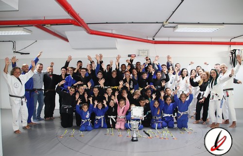 Entrenadores y niños en un dojo de judo, celebrando con una copa y medallas tras una competición, en un ambiente de alegría y triunfo, con muchos participantes y espectadores en el fondo.