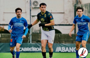 Jugadores de fútbol en un partido, dos con camiseta azul y uno con camiseta verde y blanca, en un campo de césped con fondo de muro y anuncios publicitarios.