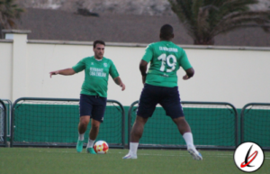 1. Dos jugadores de fútbol en entrenamiento en un campo con fondo de paredes y árboles, uno controlando el balón y el otro en posición defensiva.