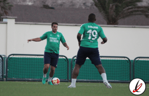 1. Dos jugadores de fútbol en entrenamiento en un campo con fondo de paredes y árboles, uno controlando el balón y el otro en posición defensiva.
