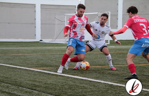 Jugador en acción peleando por el balón en un partido de fútbol en un campo artificial, con dos oponentes y un portero en el fondo.