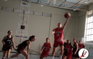 Una jugadora de baloncesto en acción lanzando el balón hacia la canasta durante un partido en un pabellón cubierto, con otras jugadoras en defensa y en ataque.