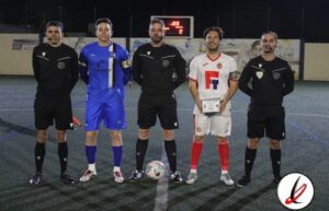 Árbitros y dos jugadores en el centro del campo de fútbol en Lanzarote, durante un partido oficial, con reloj de cronómetro y marcador visible en el fondo, en una noche de juegos.