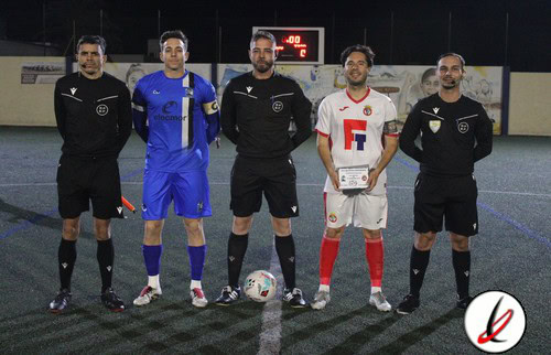 Árbitros y dos jugadores en el centro del campo de fútbol en Lanzarote, durante un partido oficial, con reloj de cronómetro y marcador visible en el fondo, en una noche de juegos.