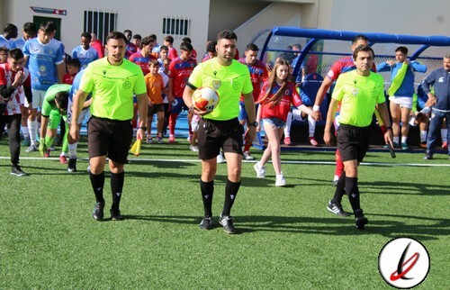Árbitros de fútbol caminando hacia el campo con niños y jugadores en el fondo en un día soleado.