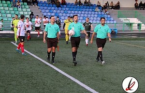 Árbitros caminando hacia el campo de fútbol antes del inicio del partido en Lanzarote Deportiva. Jugadores y espectadores en las gradas del estadio, preparado para un partido de fútbol.