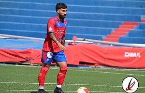 Un jugador de fútbol en el estadio durante un entrenamiento, viste uniforme rojo y azul, con un balón en el campo y asientos vacíos en las gradas.