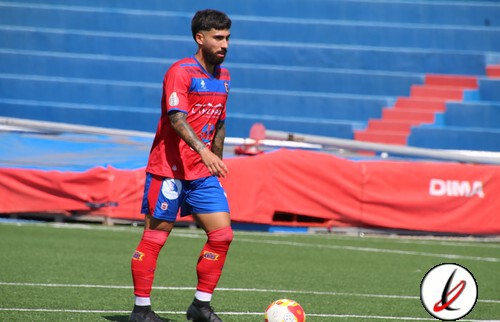 Un jugador de fútbol en el estadio durante un entrenamiento, viste uniforme rojo y azul, con un balón en el campo y asientos vacíos en las gradas.