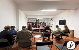 Los asistentes en una reunión o conferencia en una sala con mesas y sillas, frente a una pantalla y una máquina de premios o trofeos en una pared decorativa.