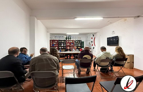 Los asistentes en una reunión o conferencia en una sala con mesas y sillas, frente a una pantalla y una máquina de premios o trofeos en una pared decorativa.