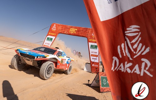 Un vehículo de rally raid atraviesa la línea de meta en el desierto durante el Rally Dakar, con banderas y arco de salida en el fondo, bajo un cielo despejado y arena en movimiento.