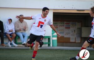 1. Jugador celebrando gol en partido de fútbol en Lanzarote Deportivo, con asistentes en banquillo y vestimenta blanca y negra.