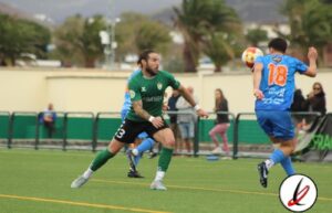 Portero en acción durante un partido de fútbol en Lanzarote deportiva, intentando bloquear un disparo del jugador con camiseta azul n°18 en un campo de césped artificial.
