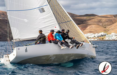 1. Grupo de cinco personas navegando en un velero de regata en el océano, con montañas y pueblo en el fondo, día soleado y viento moderado.