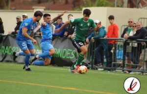 1. Jugador en verde controlando el balón durante un partido de fútbol en Lanzarote, con defensores en azul y público en las gradas.