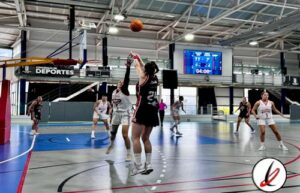 Juga de baloncesto femenino en una cancha cubierta, con jugadoras en movimiento, una de ellas saltando para bloquear un tiro, en un partido con marcador digital y público en las gradas.