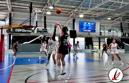 Juga de baloncesto femenino en una cancha cubierta, con jugadoras en movimiento, una de ellas saltando para bloquear un tiro, en un partido con marcador digital y público en las gradas.