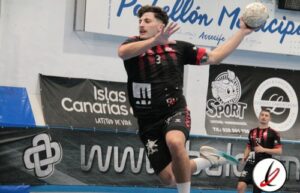 Atleta de handball en acción lanzando una pelota en un partido en interior, con jugadores en el fondo y publicidad en las paredes del gimnasio.