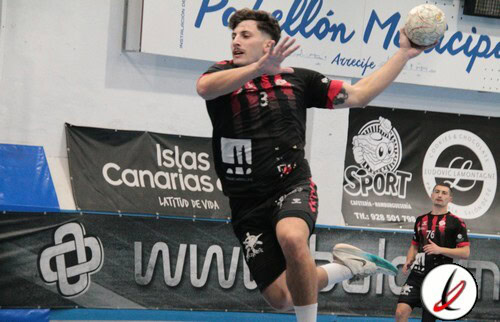 Atleta de handball en acción lanzando una pelota en un partido en interior, con jugadores en el fondo y publicidad en las paredes del gimnasio.