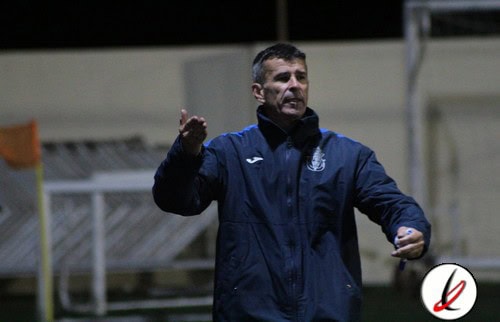 Entrenador en el campo durante un partido de fútbol en Lanzarote, vistiendo chaqueta deportiva azul y gesticulando con la mano, en un entorno de estadio o campo de juego.