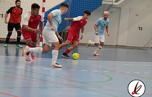 Jugadores de fútbol sala en acción durante un partido en pista cubierta con fondo de red y paredes blancas.