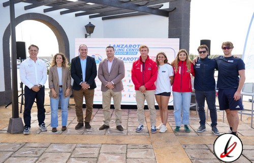 1. Grupo de siete personas, incluyendo atletas y oficiales, posando en un evento deportivo en Lanzarote, con vista al mar y un escenario con carteles y altavoces en el fondo.
