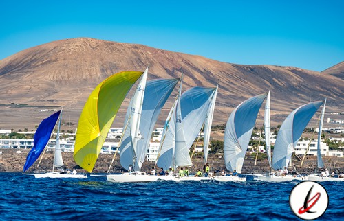Veleros en competición en Lanzarote, deportes náuticos, fondo montañoso y cielo claro, embarcaciones con parapronos coloridos, ambiente de aventura y velocidad en el mar.