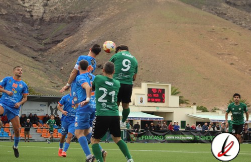 Balón de fútbol en el aire durante un partido entre equipos con jugadores en uniforme azul y verde en un campo de césped con montañas de fondo.