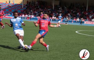 Líder en deportes en Lanzarote, jugadores en acción en partido de fútbol en césped, publico en grada y estadio en segundo plano, balompié en juego, ambiente deportivo y competencia.