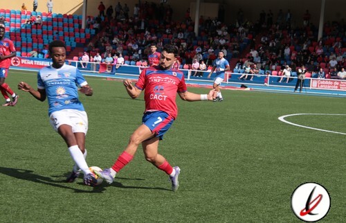Líder en deportes en Lanzarote, jugadores en acción en partido de fútbol en césped, publico en grada y estadio en segundo plano, balompié en juego, ambiente deportivo y competencia.