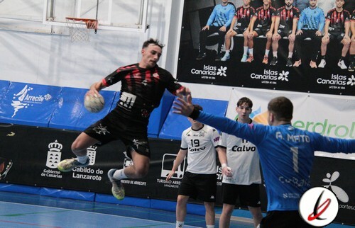 Lanzador de balonmano en acción lanzando contra el portero en un partido en Lanzarote. Jugadores en el campo, con un cartel de Islas Canarias y un fondo de instalaciones deportivas cubiertas.