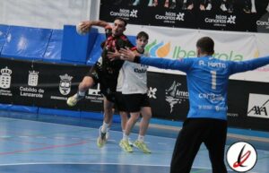 Jugadores de balonmano en acción en la cancha de un estadio cubierto, con el portero en posición defendiendo y dos jugadores en movimiento durante un partido en Lanzarote.
