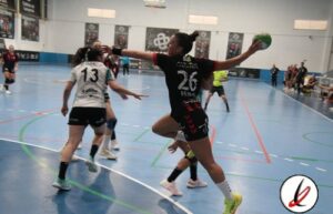 Lanzadora de balonmano en acción durante un partido femenino en un pabellón cubierto, con jugadoras en movimiento y público en las gradas.