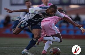 Un jugador con camiseta azul número 25 lucha por el balón con un jugador con camiseta rosa en un campo de fútbol. Ambos intentan ganar posesión mientras mantienen un esfuerzo intenso durante el partido.