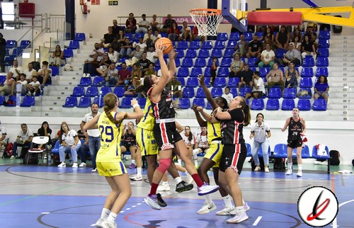 Un equipo de baloncesto femenino en acción durante un partido en una cancha cubierta con público en las gradas. Jugadoras en movimiento intentan anotar mientras la defensa trata de bloquear el tiro.