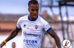 Atlético de Lanzarote jugador en acción durante un partido de fútbol en el campo del equipo, vistiendo camiseta blanca con patrocinadores y logotipos, concentrado en el juego.