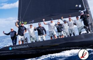 Nave de regata con equipo de vela celebrando en el mar, con bandera negra y logotipo de Peninsula, en un día soleado con cielo despejado.