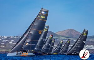 Velero de regata con varias embarcaciones en competición en Lanzarote, fondo de montañas y cielo despejado.