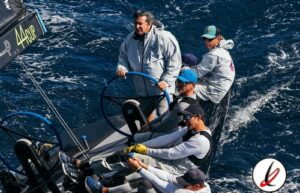Navegantes en un barco de regata en el océano, con mar agitado y viento fuerte, cuatro personas con ropa de protección y gafas, controlando la embarcación durante una competición náutica.