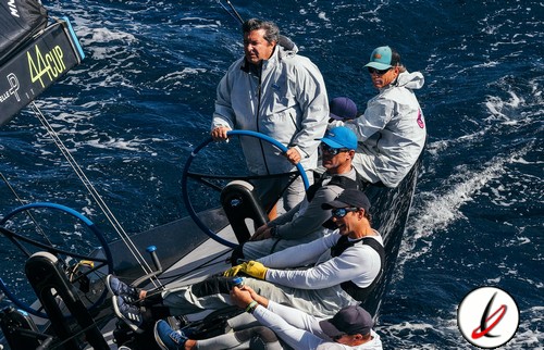 Navegantes en un barco de regata en el océano, con mar agitado y viento fuerte, cuatro personas con ropa de protección y gafas, controlando la embarcación durante una competición náutica.