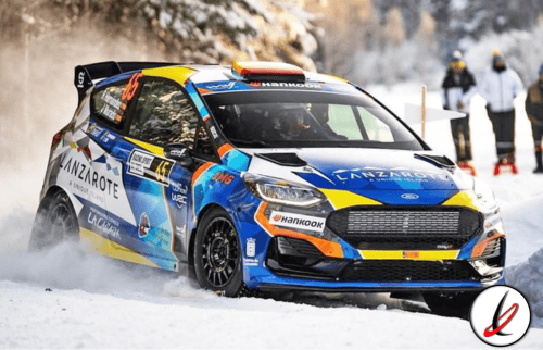 Coche de rally en nieve, piloto en acción en un entorno invernal con espectadores en el fondo.