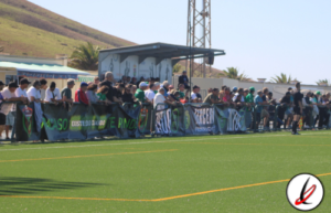Jugadores y espectadores en la grada de un campo de fútbol en Lanzarote, con montañas y palmeras al fondo, durante un partido o evento deportivo.